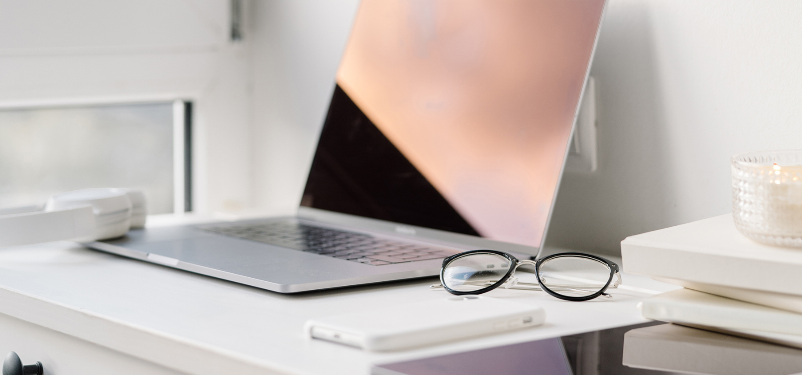 A modern laptop on a white desk with glasses, a smartphone, and headphones nearby, illuminated by natural light.